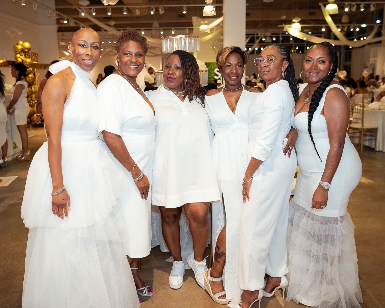 A group in white dresses poses together at an elegant indoor celebration.