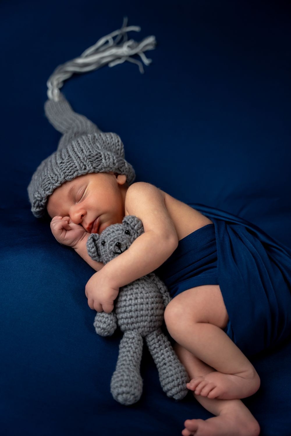 Sleeping newborn baby in navy blue swaddle cuddles with gray knitted teddy bear against dark background.