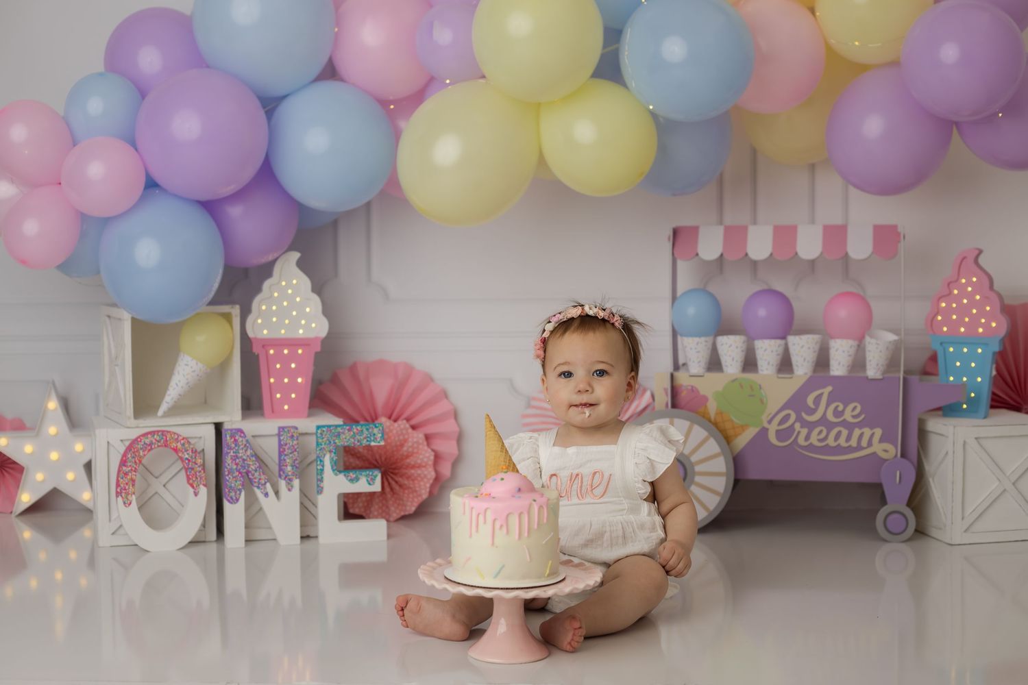 An adorable baby girl sitting with a cake in her custom pastel ice-cream theme background for her studio cake smash session.