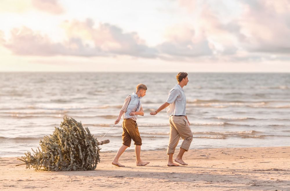 Father and son walking barefoot on beach carrying Christmas tree at sunset.