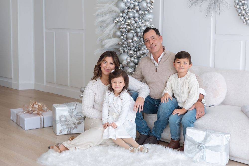 Family of four sitting together near silver Christmas tree with wrapped gifts.