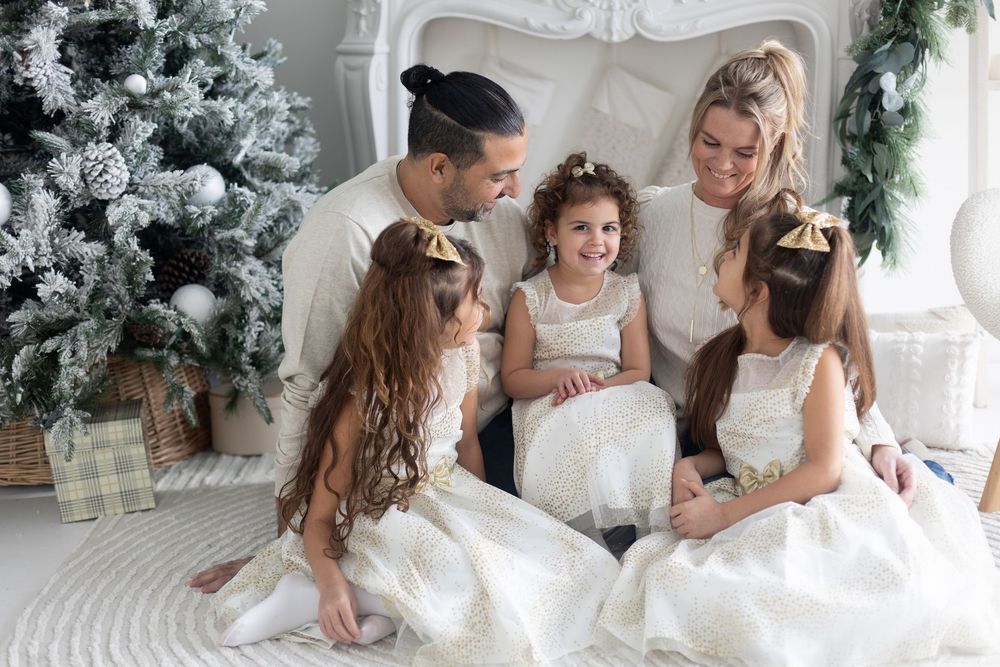Parents with three daughters in white dresses gathered around Christmas tree.