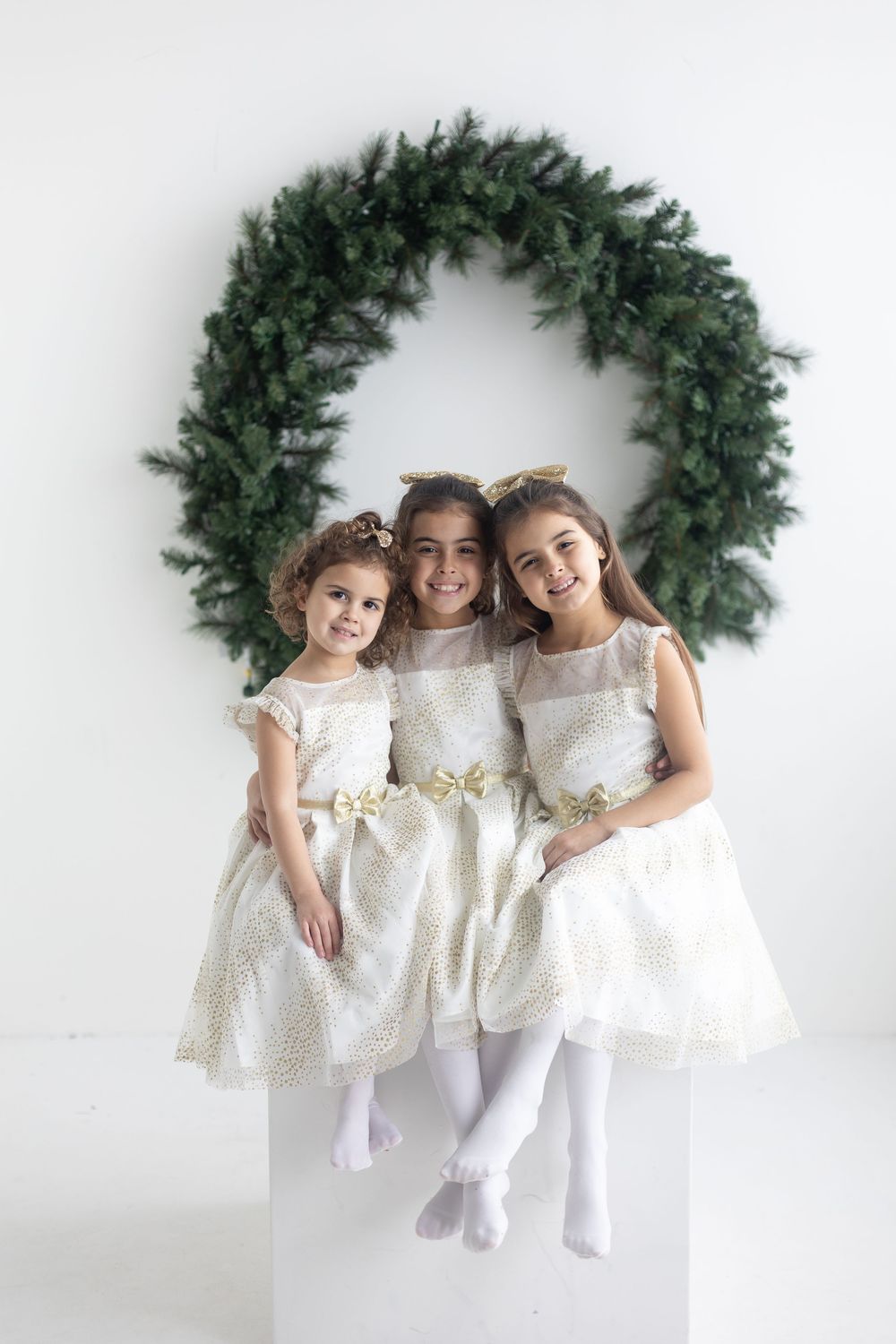 Three girls in gold and white dresses posing before large green wreath.