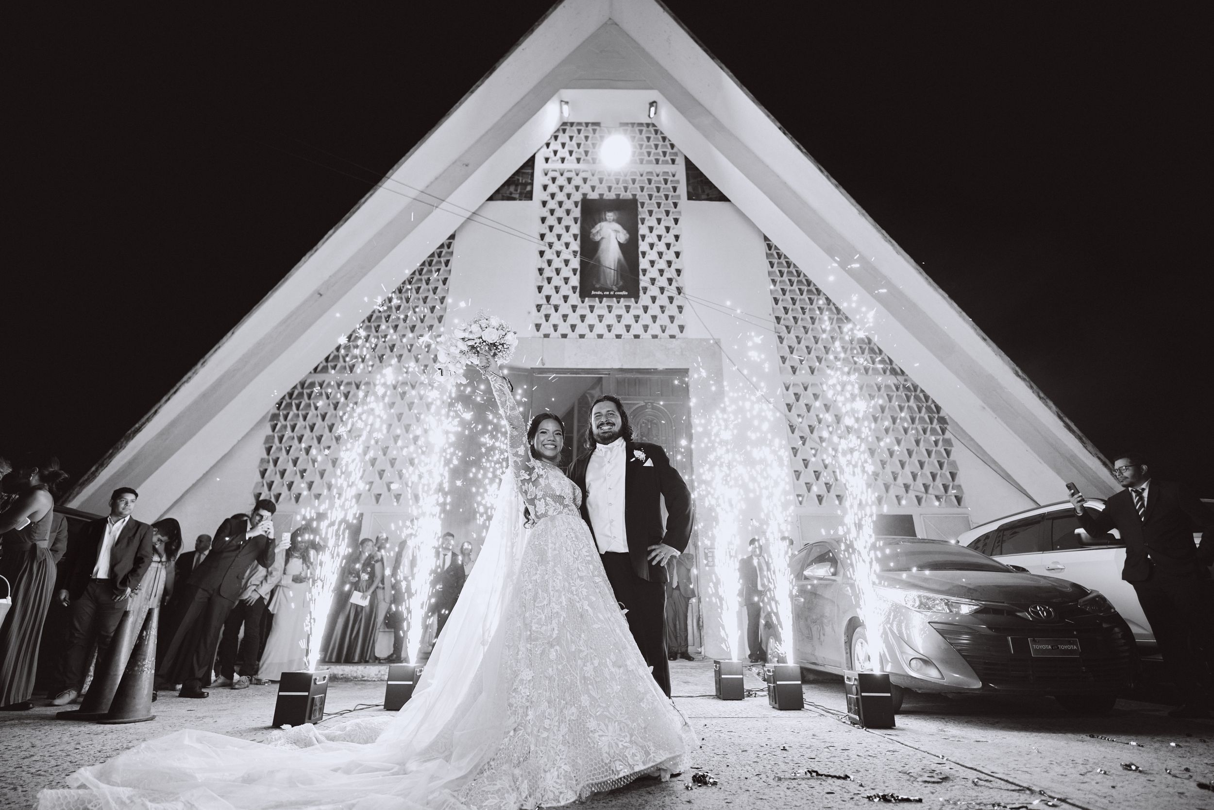 A dramatic black and white photo of a wedding couple in front of an illuminated chapel.