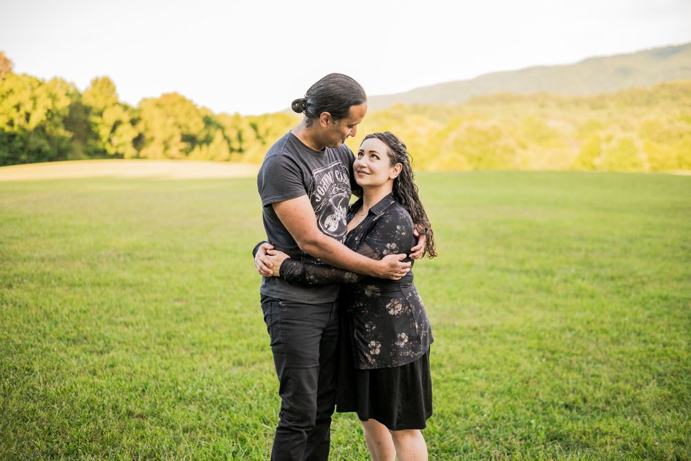 Hispanic couple embracing and smiling in a green field with mountains and golden trees in the background at sunset. Couples Photographer.