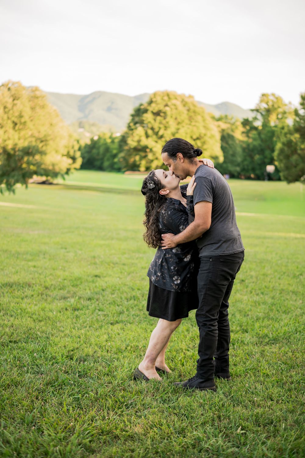 Hispanic Couple sharing a romantic kiss in a lush park with mountains and trees in the background at sunset. Couples Photographer.