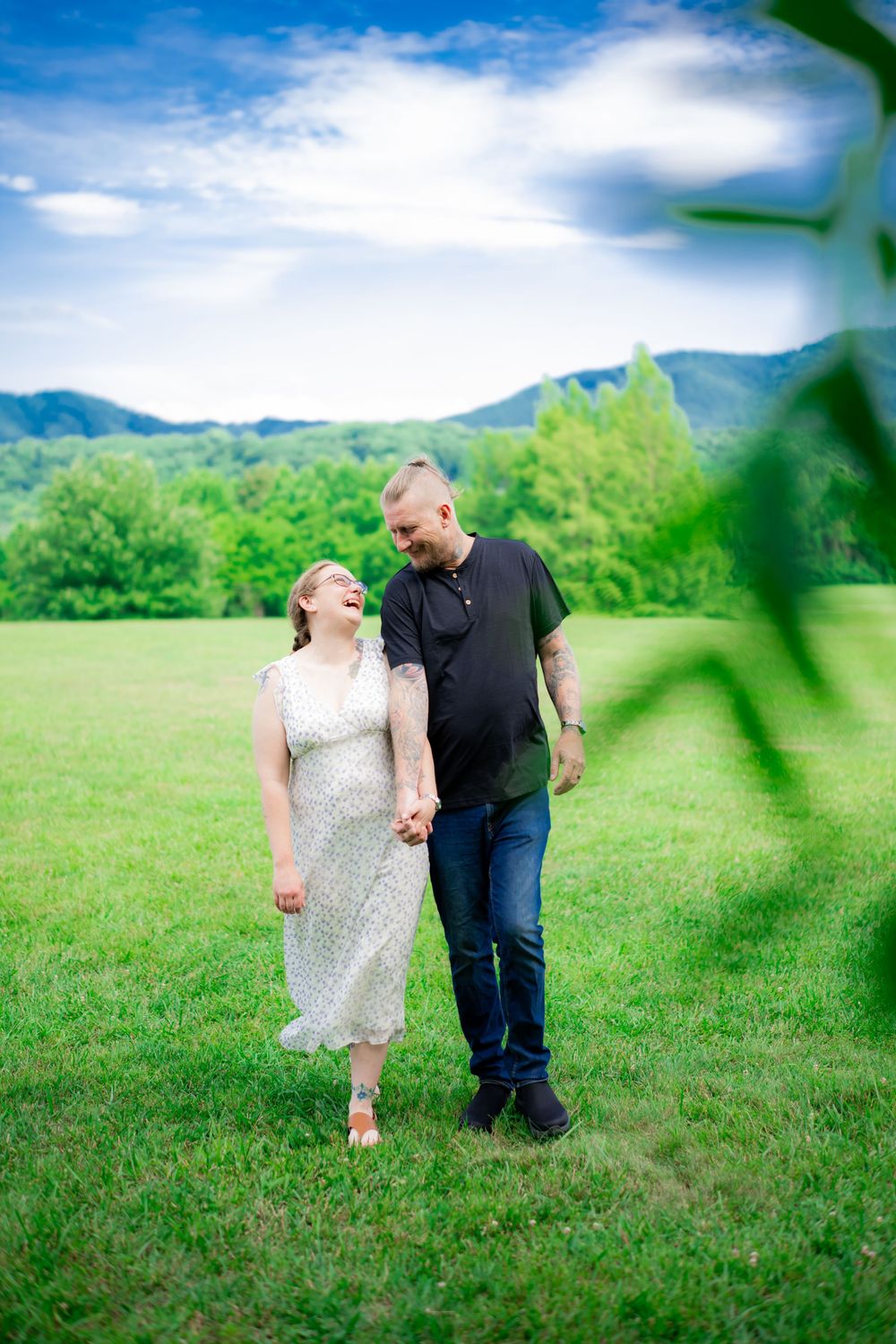 A laughing couple walks hand-in-hand through a green meadow with mountain views under a blue sky. Engagement Photographer.