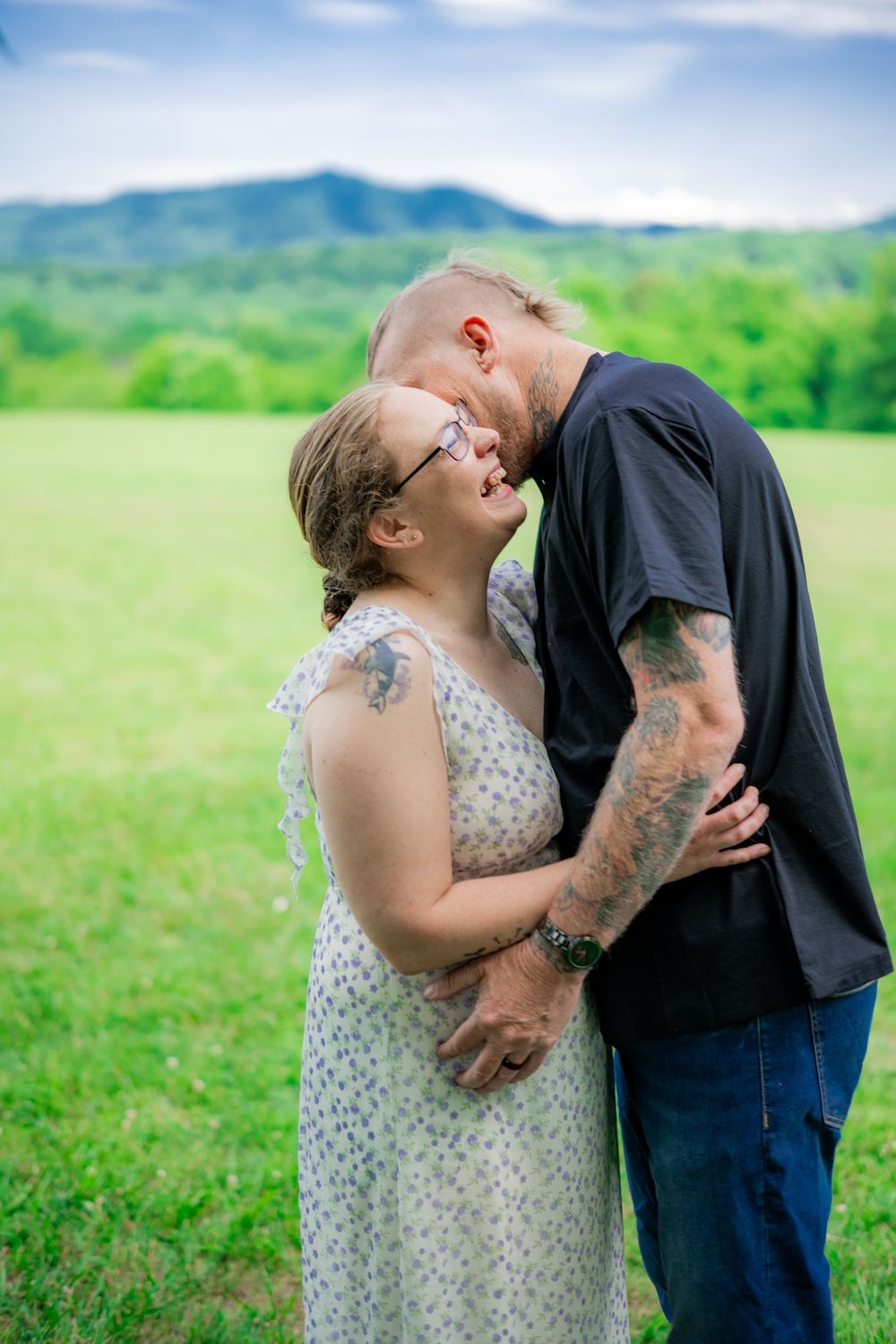 Couple with tattoos sharing a romantic moment outdoors, embracing in a green field with mountains in the background. Engagement Photographer