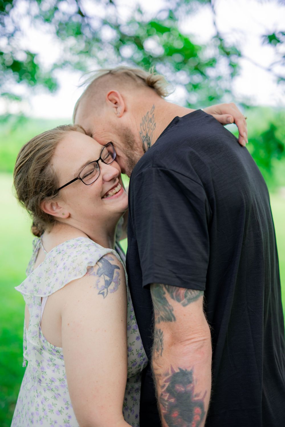Couple sharing a tender moment outdoors, woman smiling in floral dress and glasses, man in black t-shirt. Engagement Photographer.