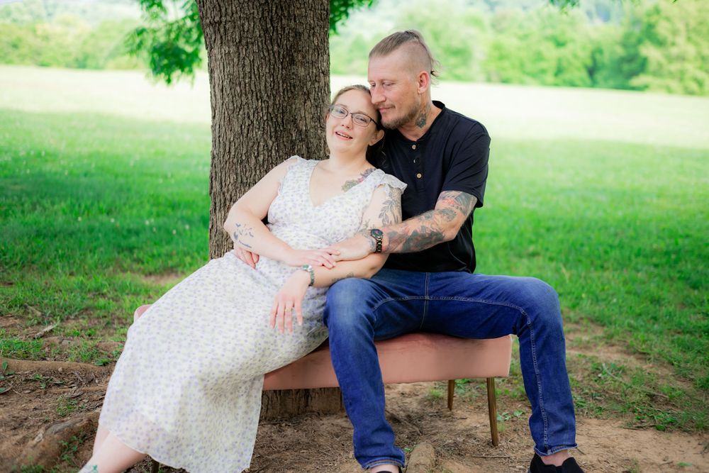 Couple sitting together under a tree on a bench in a park, woman in floral dress leaning on man in black shirt. Engagement Photographer.
