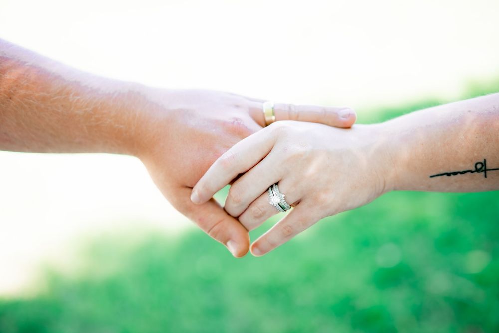 Two hands holding, both wearing wedding rings, with a green bokeh background outdoors. Couples, Engagement Photographer.