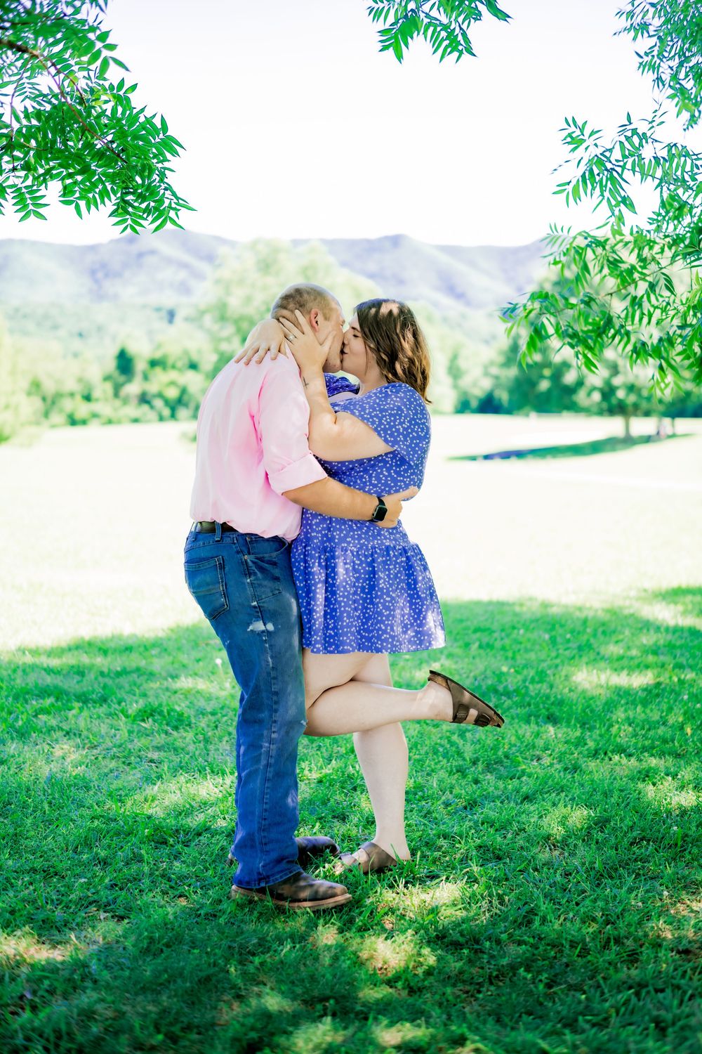 Couple sharing a romantic kiss outdoors on green grass with mountain views and lush trees in background. Couples Photographer.