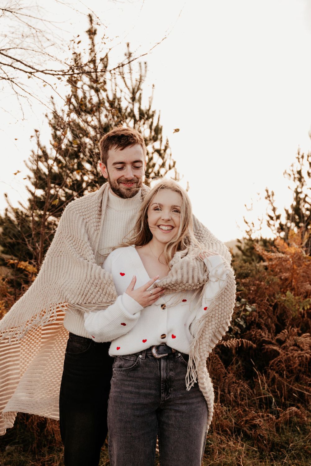 Happy couple in warm sweaters sharing a tender moment against pine trees and fall foliage.