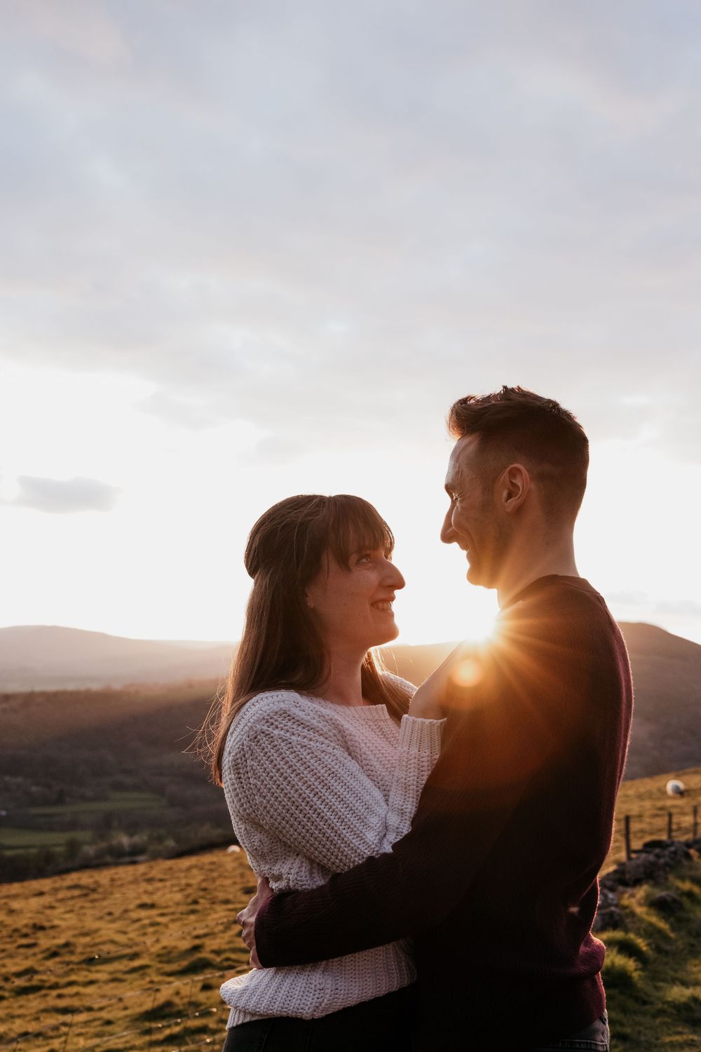 A couple embraces at sunset on a hilltop, sharing a romantic moment with golden light and rolling mountains behind them.