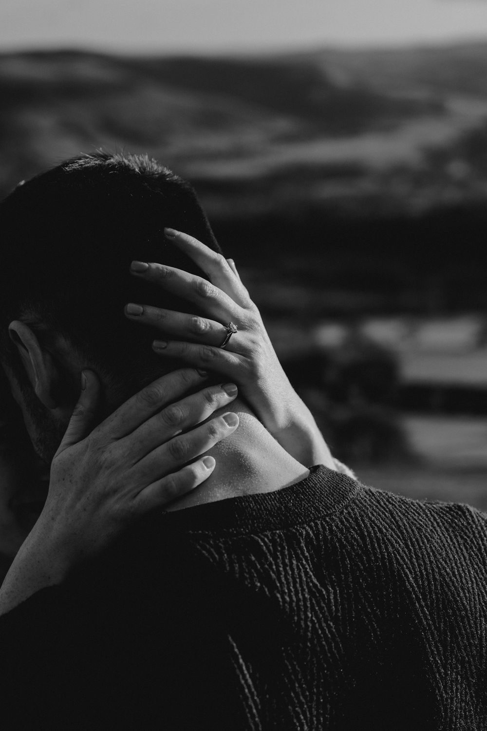Black and white photo of a person holding their head in their hands outdoors with rolling hills in the background.