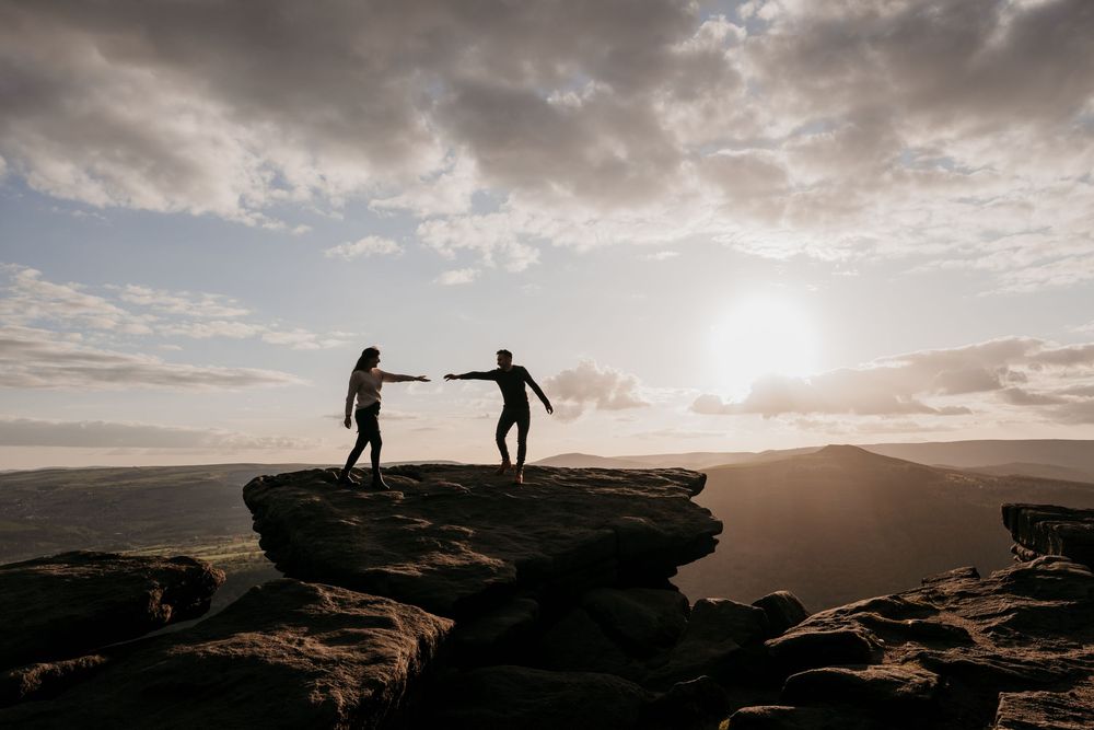 Two silhouetted figures holding hands on a rocky summit at sunset with dramatic cloudy skies and rolling hills behind.