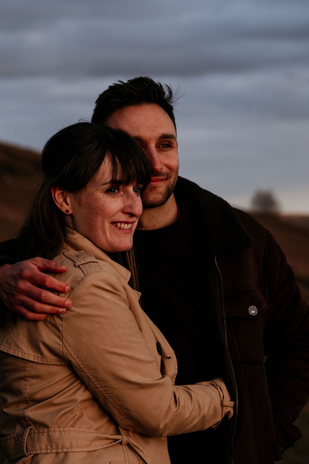 A smiling couple embraces outdoors at golden hour, woman in beige jacket, man in black, with cloudy sky behind them.