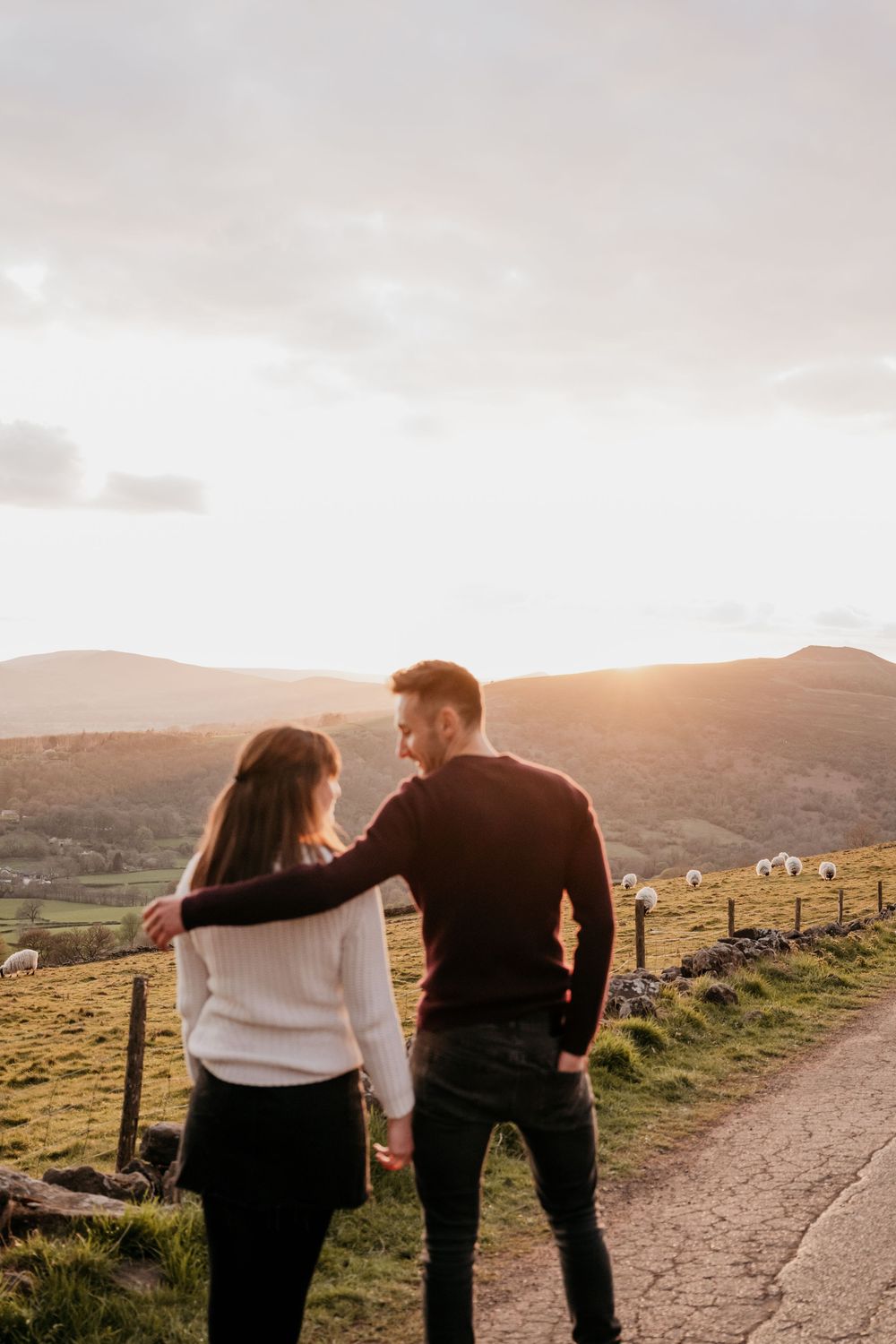 Couple embracing on a scenic mountain path at golden sunset, with rolling hills and grazing sheep in the background.