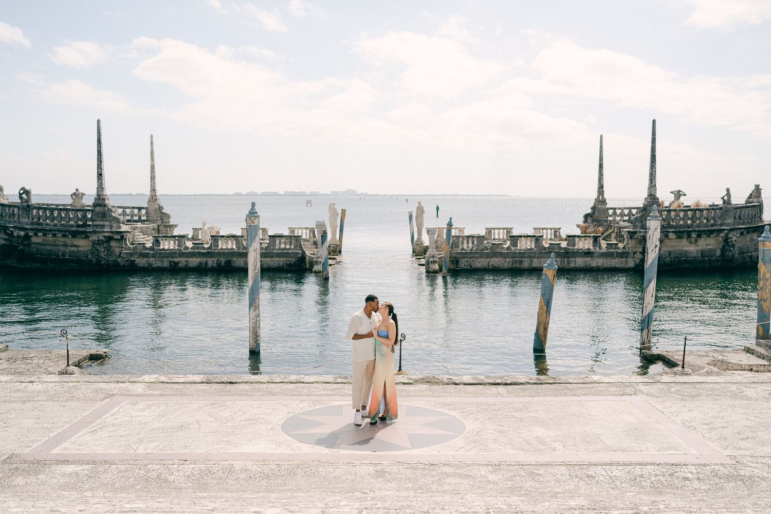 Couple embracing on Vizcaya Museum and Gardens with ornate stone pillars and calm blue water in the background.