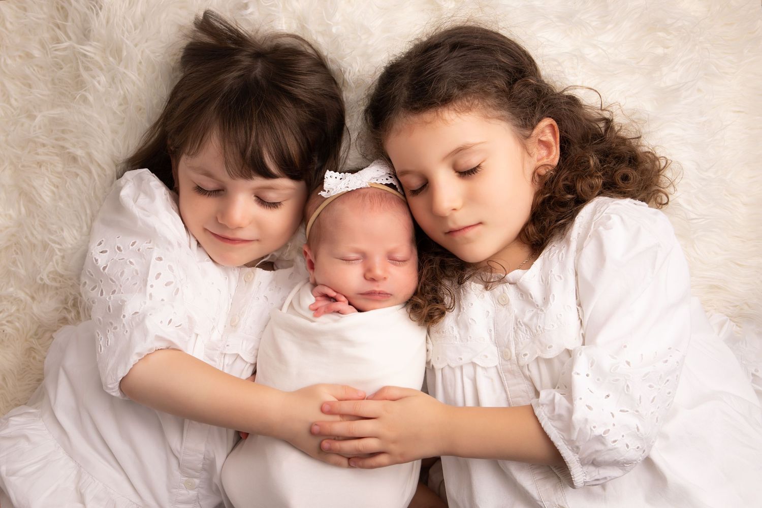 Children in white dresses gather around sleeping newborn for studio portrait.