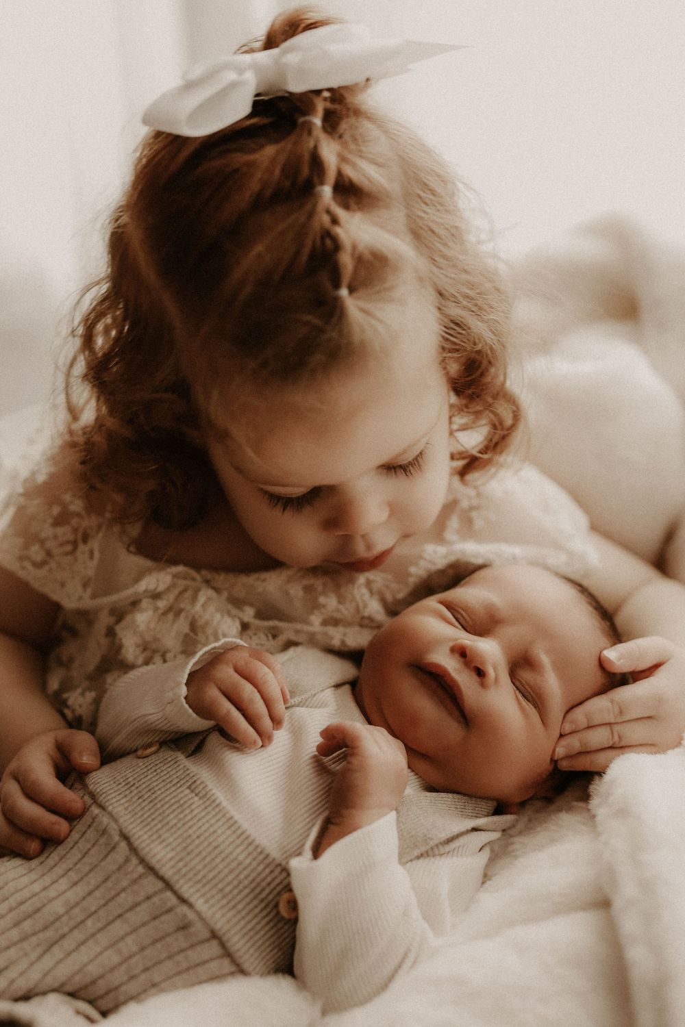 Toddler girl with white bow gently kisses sleeping newborn baby sibling in a soft, warm portrait.