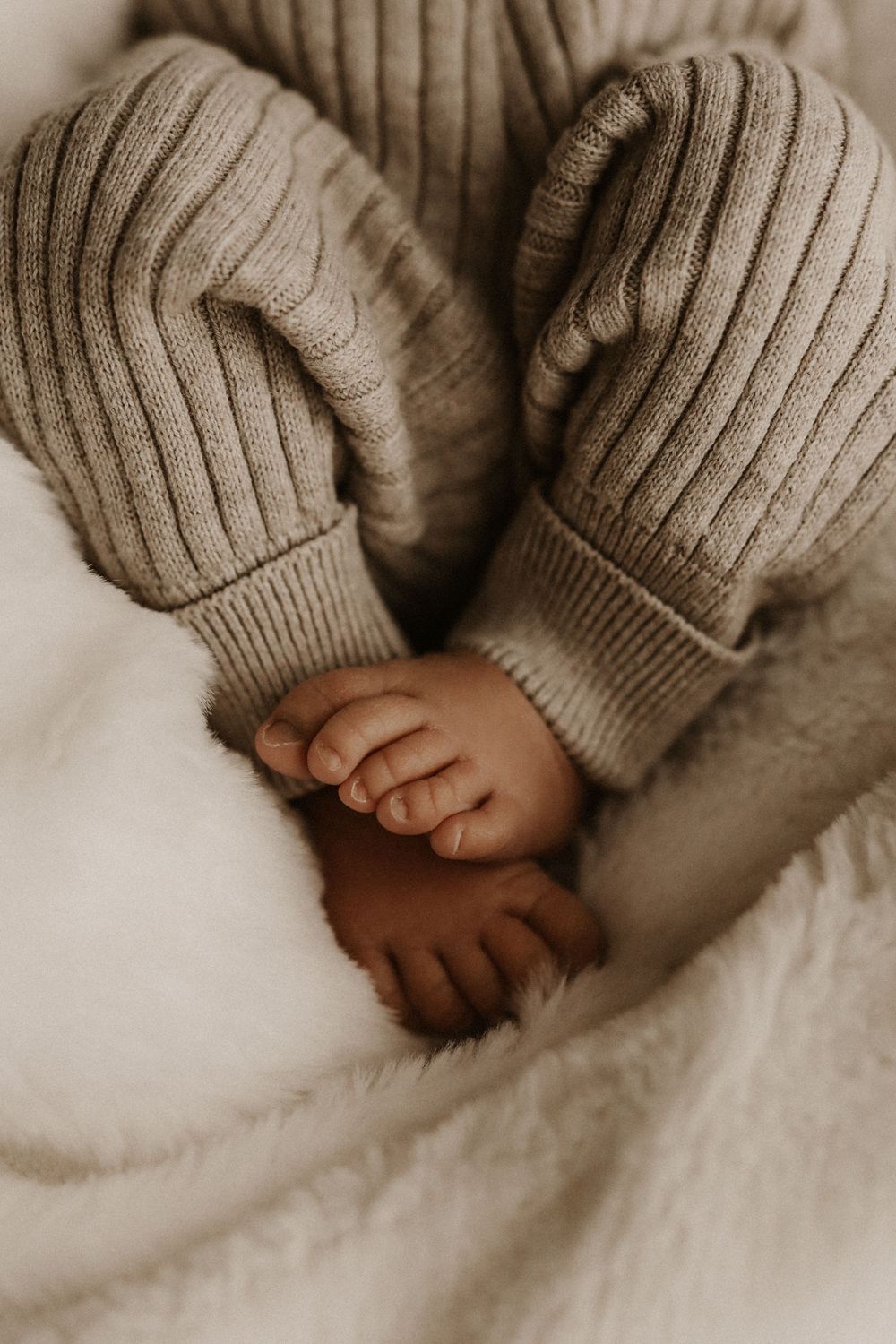 Close-up of a baby's tiny hands and feet in a ribbed knit beige romper, resting on a soft white blanket.