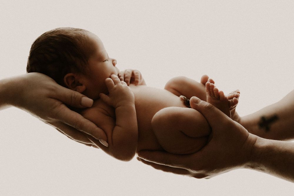 Newborn baby cradled gently in two adult hands against a soft white background.