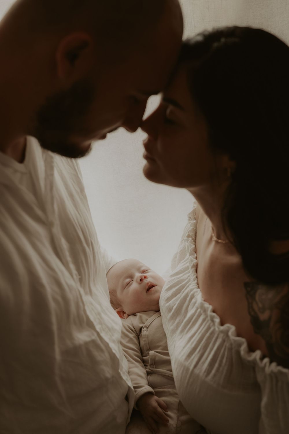 A couple tenderly touches foreheads while cradling their newborn baby, captured in a warm, intimate family portrait.