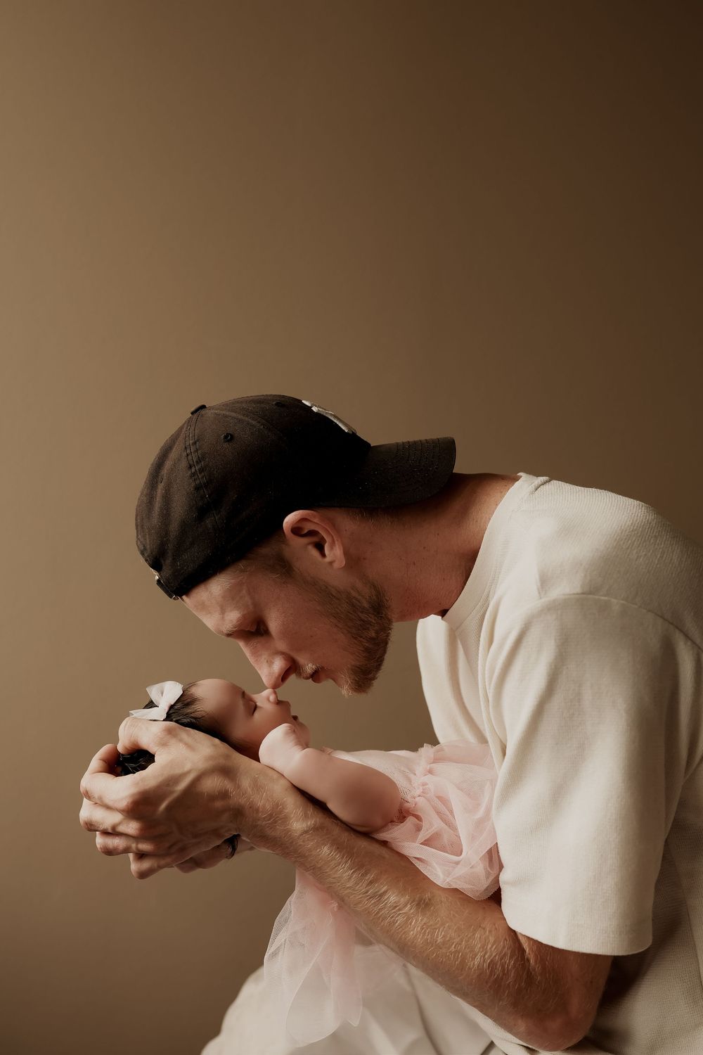A bearded father in a cap tenderly kisses his newborn baby dressed in pink, against a warm beige background.