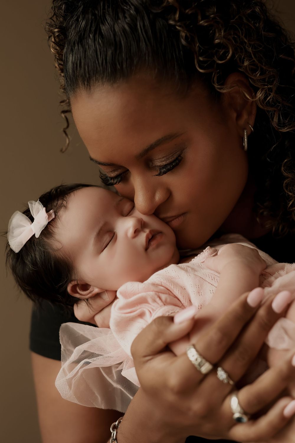 A mother tenderly kisses her sleeping newborn baby dressed in pink, captured in a warm portrait.