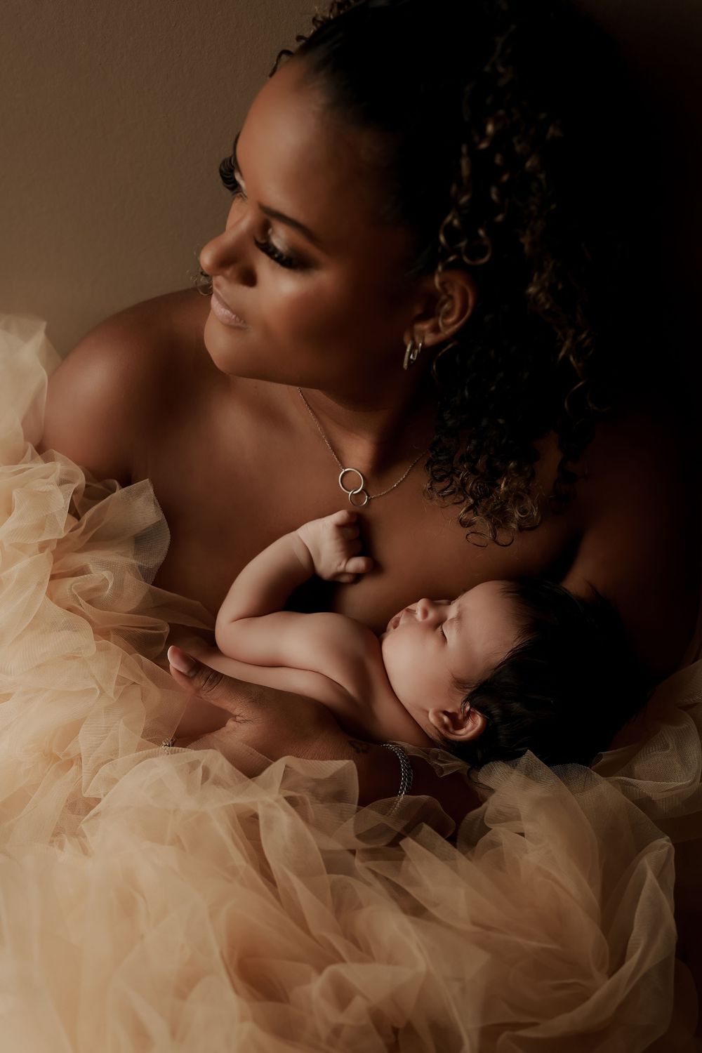 A mother in a beige tulle gown tenderly holds her sleeping newborn baby in a warm, softly lit portrait.