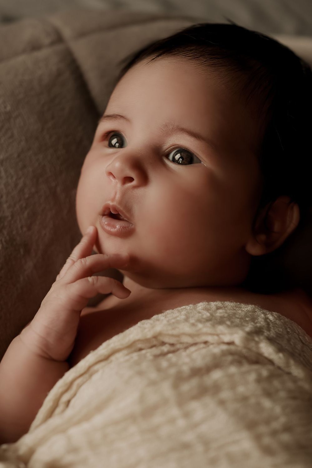 Adorable baby with dark hair and wide eyes lying on a soft blanket, finger resting on chin.