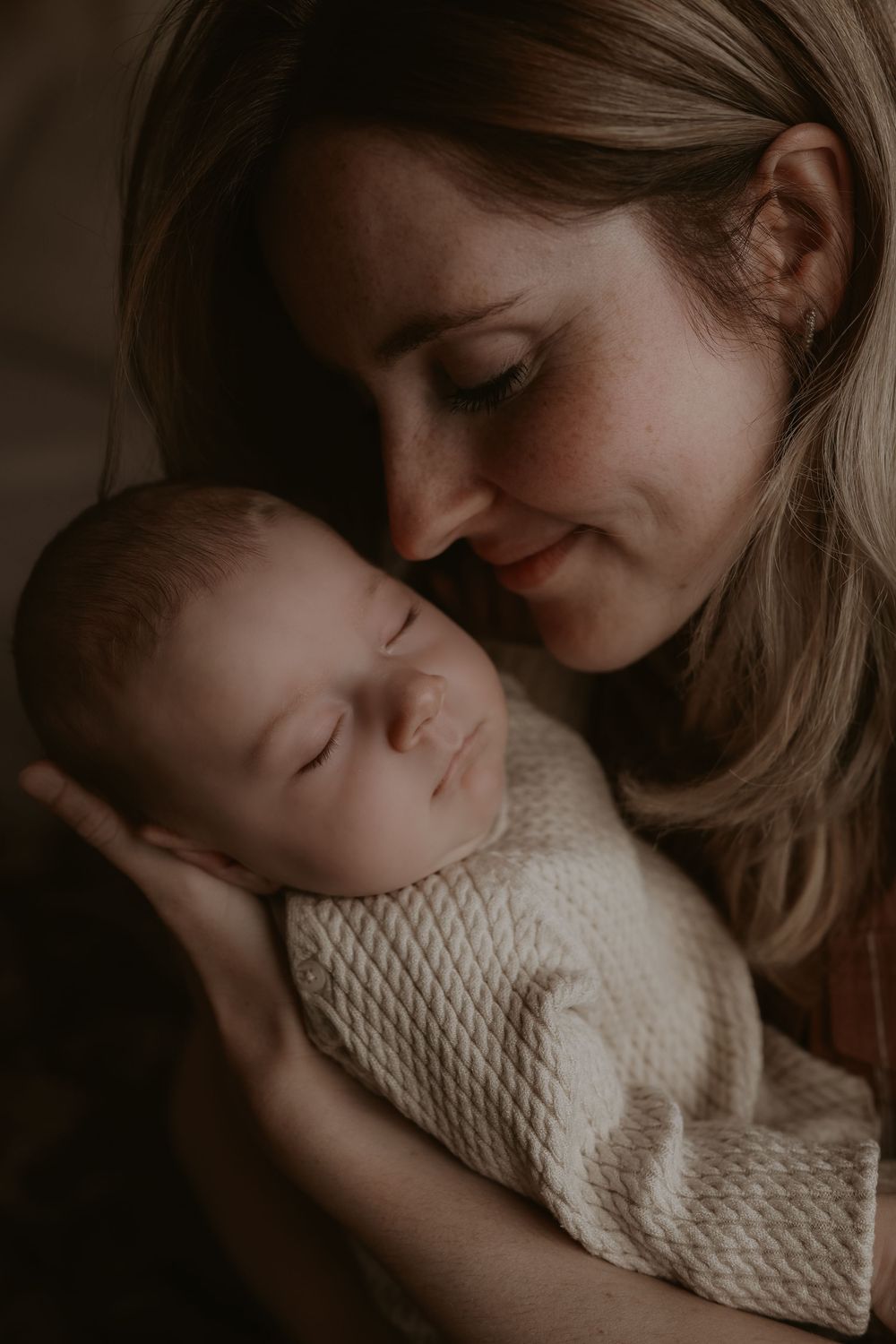 A smiling mother tenderly holds her sleeping newborn baby wrapped in a soft white knit blanket.
