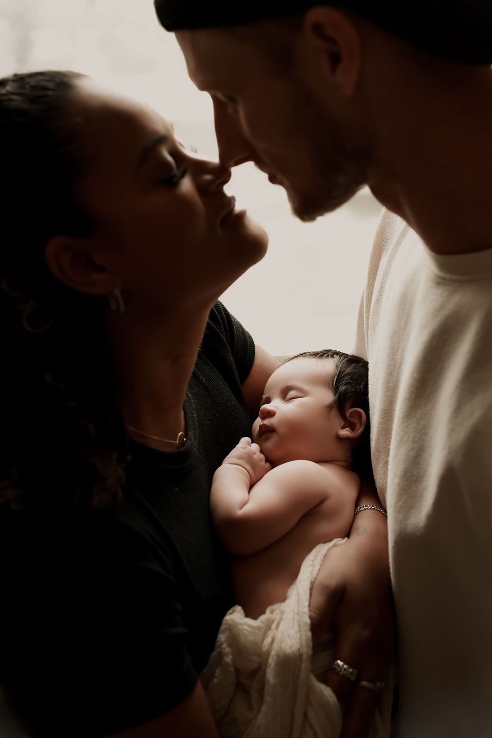 Couple tenderly touching noses while cradling their sleeping newborn baby in a softly lit intimate portrait.
