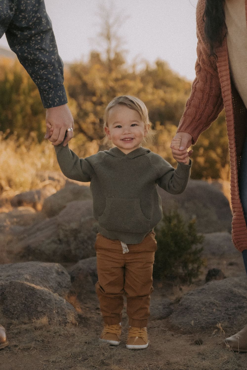 A small child in a green sweater and brown pants holds hands with adults while walking on rocky terrain at sunset in the Sandia foothills.