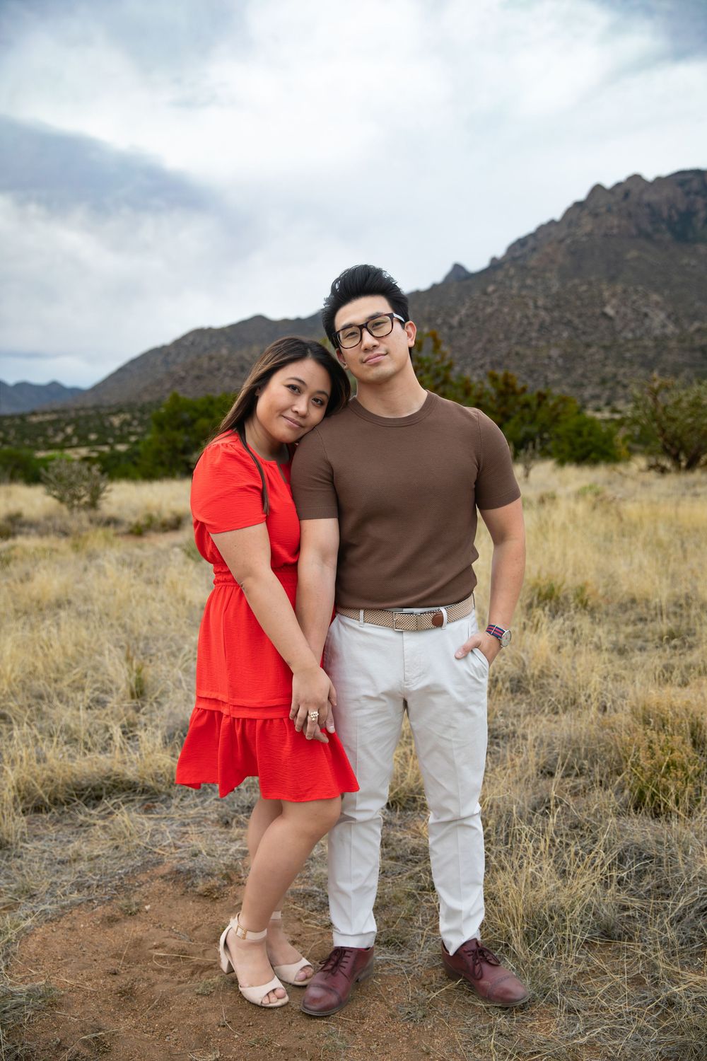 Couple posing in desert landscape with mountains, woman in red dress leaning on man in brown shirt and white pants.