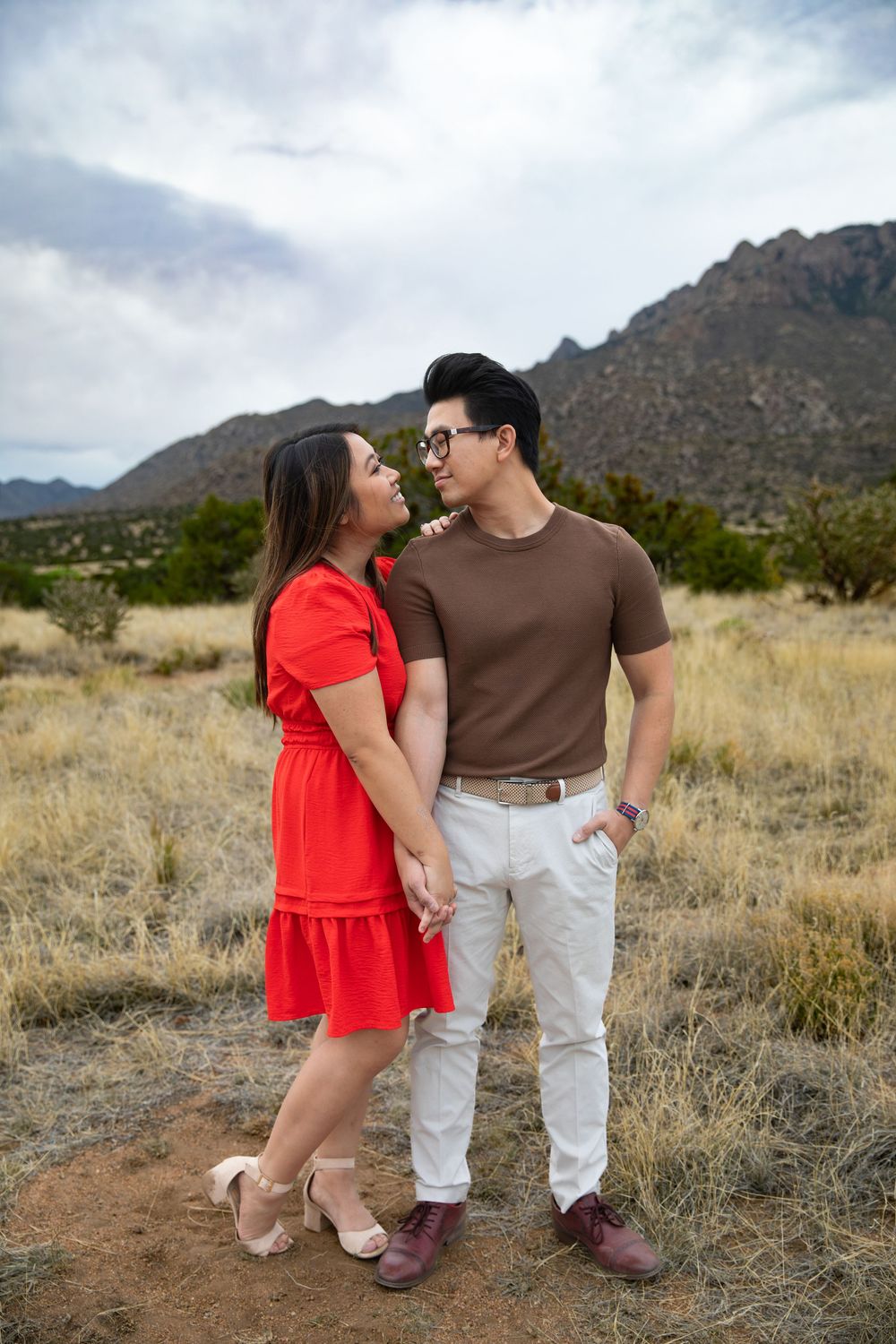A couple holds hands and gazes at each other in a desert landscape with mountains in the background.