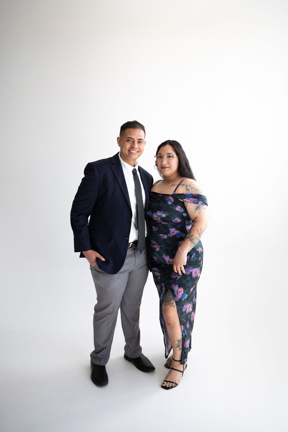 Couple posing together in formal attire against white background, man in navy blazer and woman in floral off-shoulder dress.