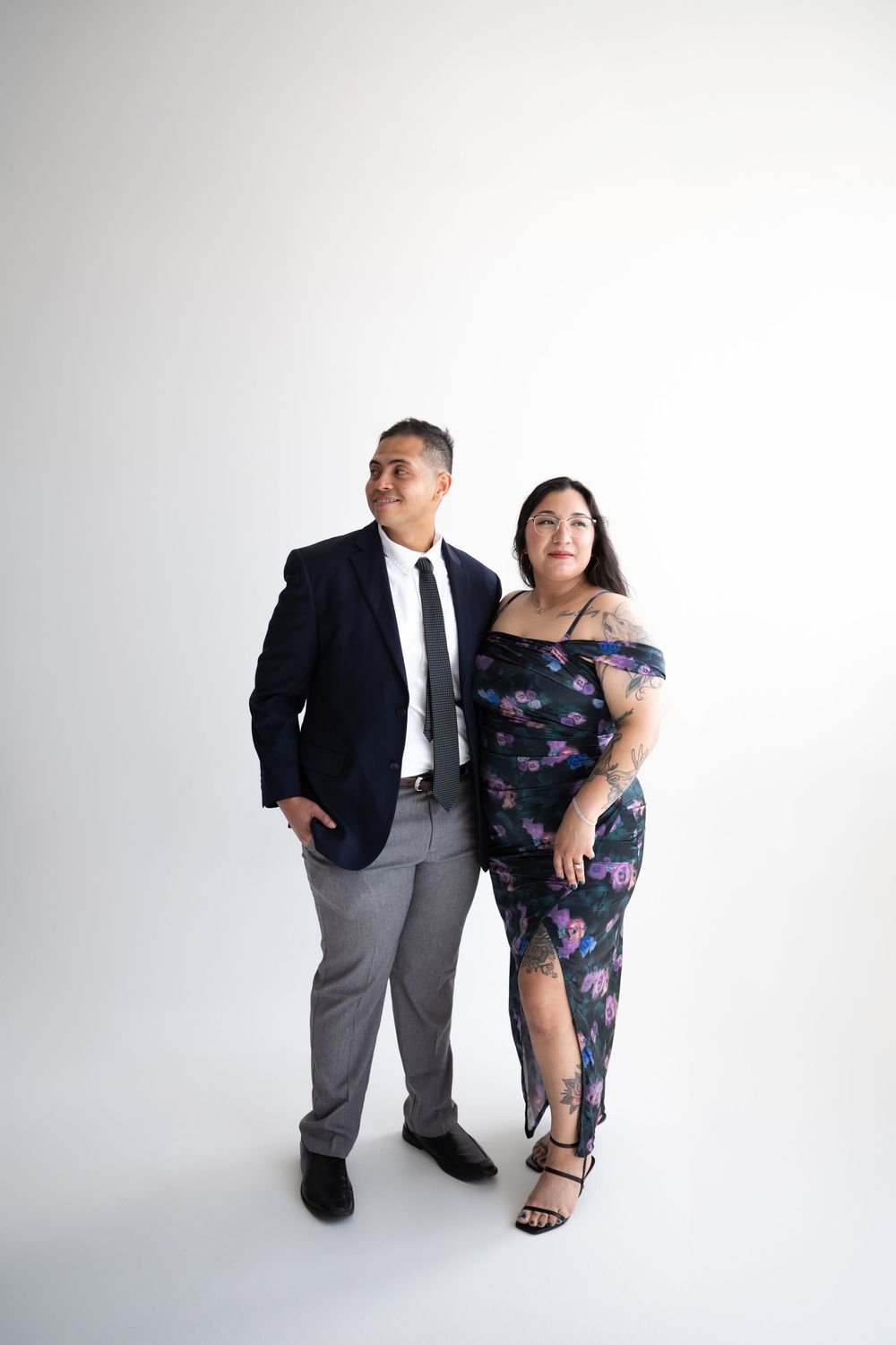 A couple poses formally, man in navy suit and woman in floral off-shoulder dress, against white background.