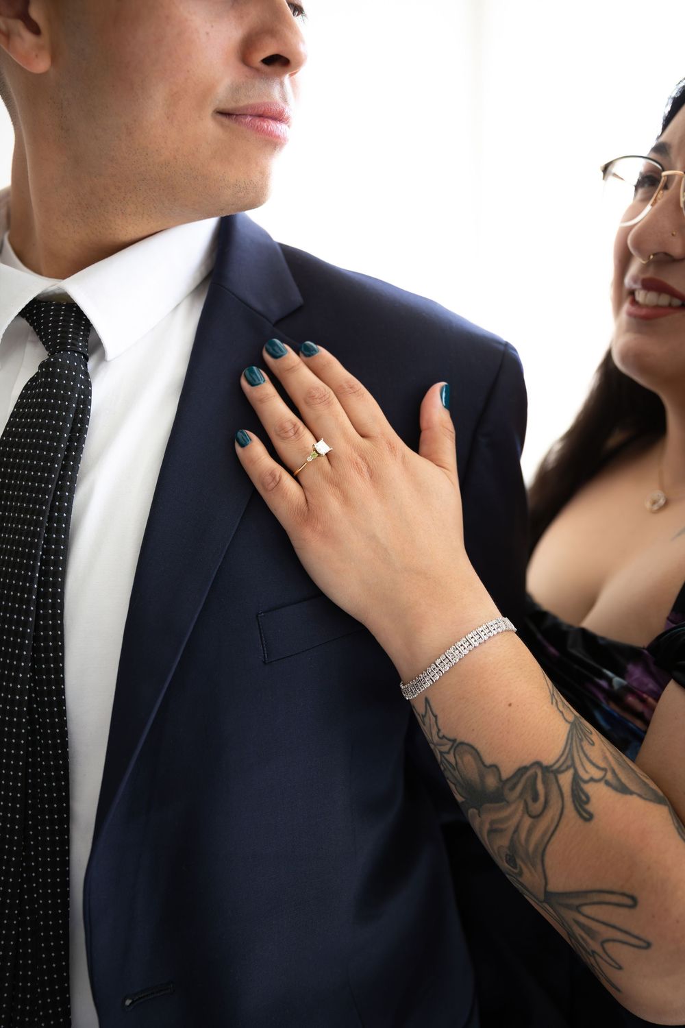 Close-up of woman's hand with engagement ring and teal nails resting on man's navy suit jacket with black tie.