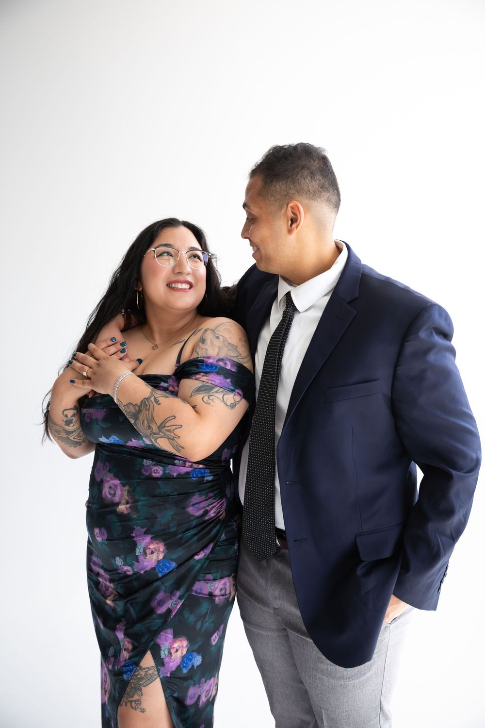 A couple poses together, woman in floral dress with tattoos and man in navy suit with black tie, against white background.