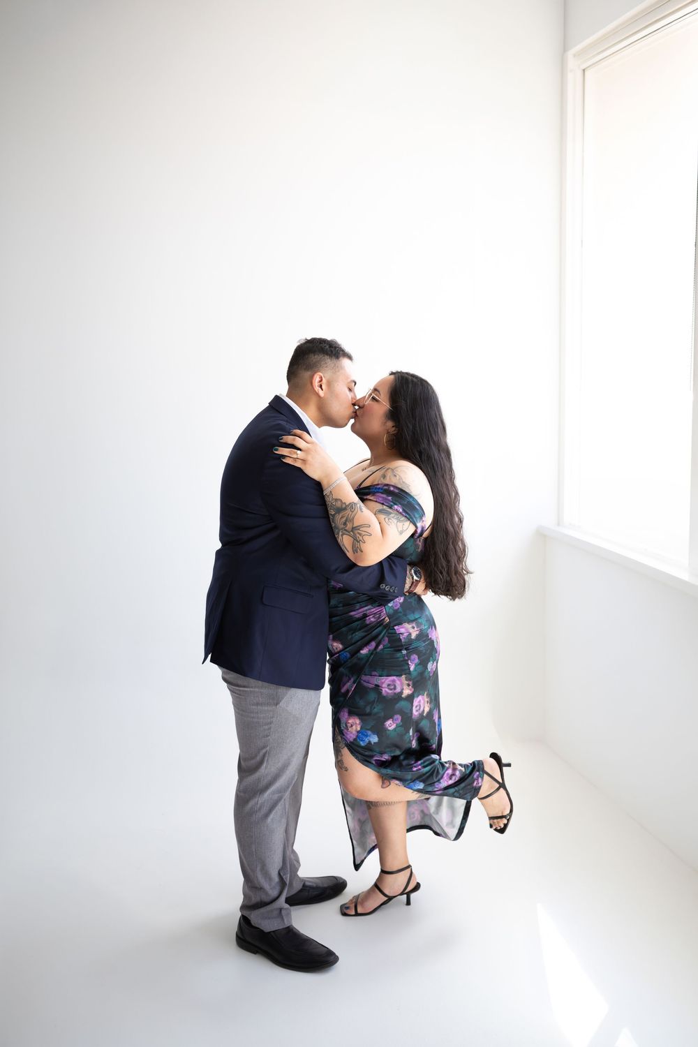 A couple shares a romantic kiss in a bright white studio, the man in a navy blazer and the woman in a floral dress.