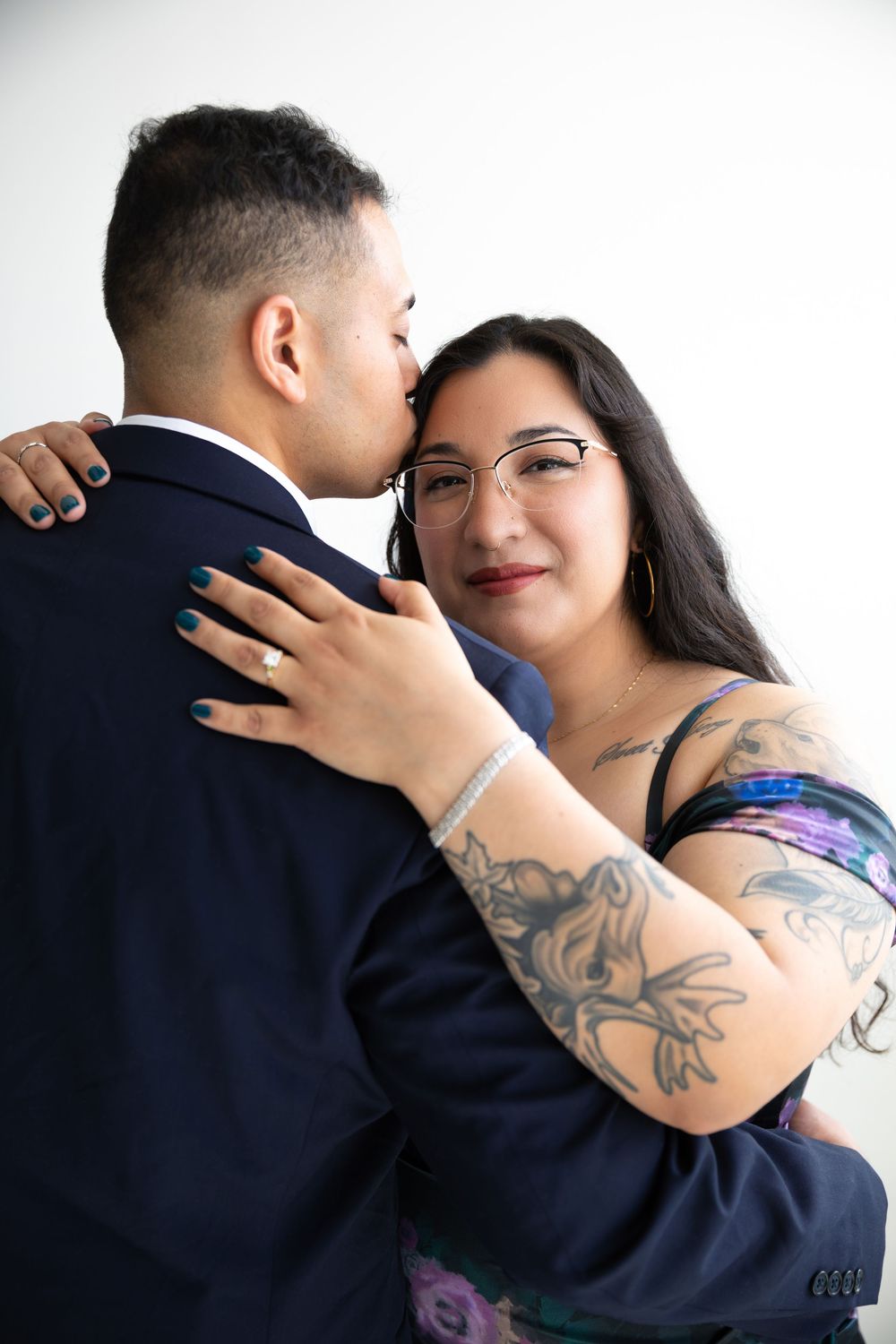 Engaged couple embracing closely, woman with tattoos and glasses wearing floral dress showing diamond ring, man in navy suit.