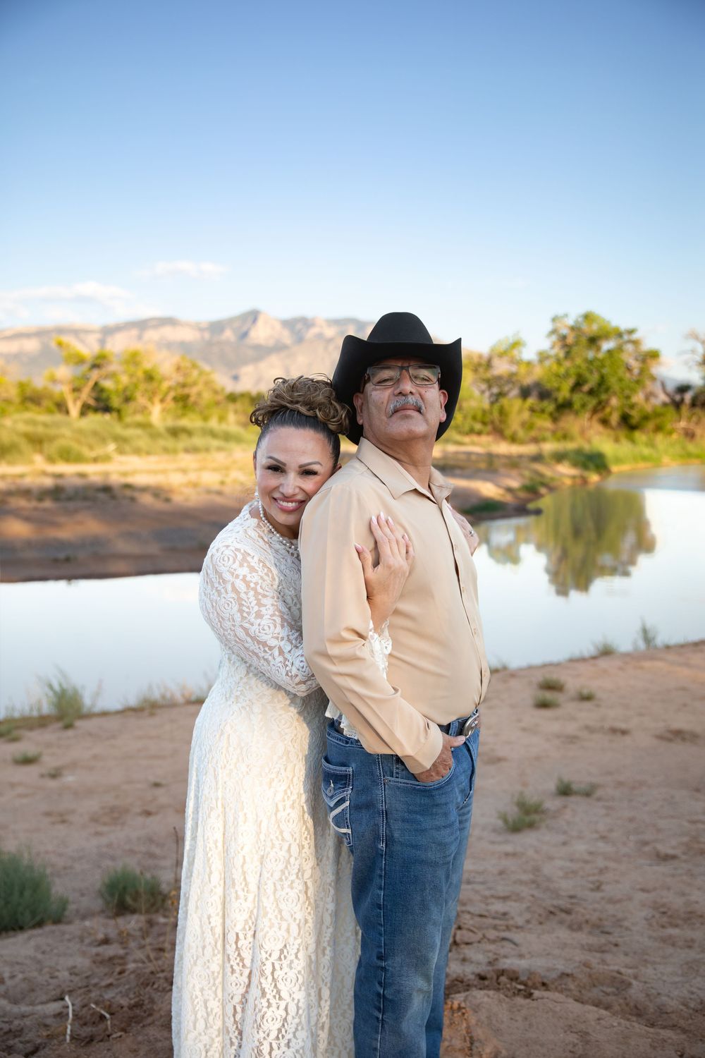 Bride in white lace dress embraces groom in cowboy hat and jeans near a scenic river with mountains at sunset.