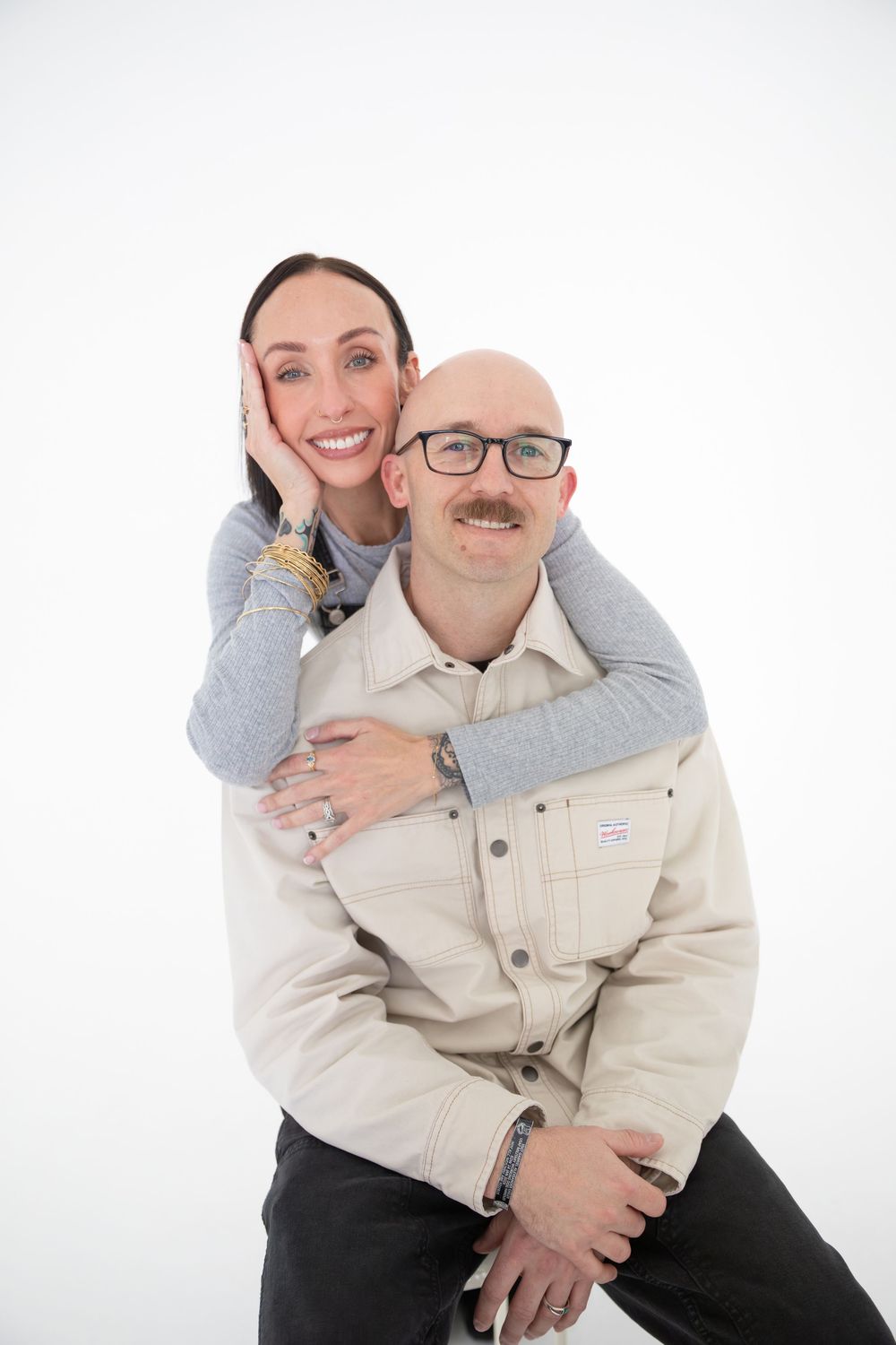 A couple poses together against a white background, with one person hugging the other from behind in a playful embrace.