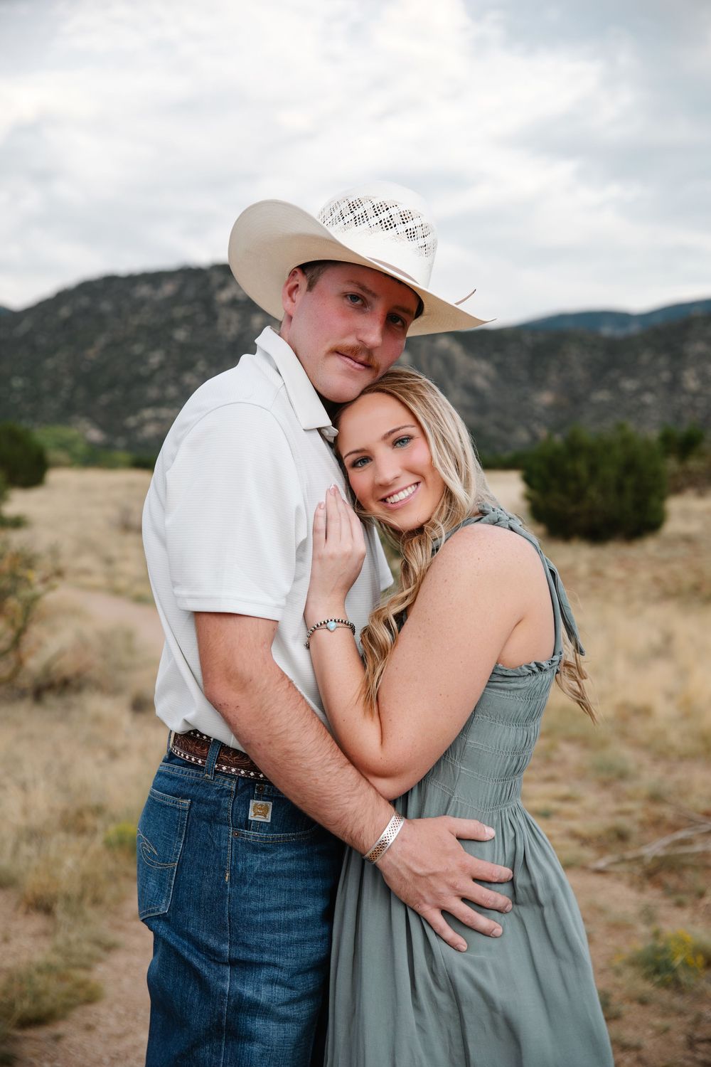 Couple embracing in rustic desert landscape, man in cowboy hat and jeans, woman in sage green dress, mountains behind.
