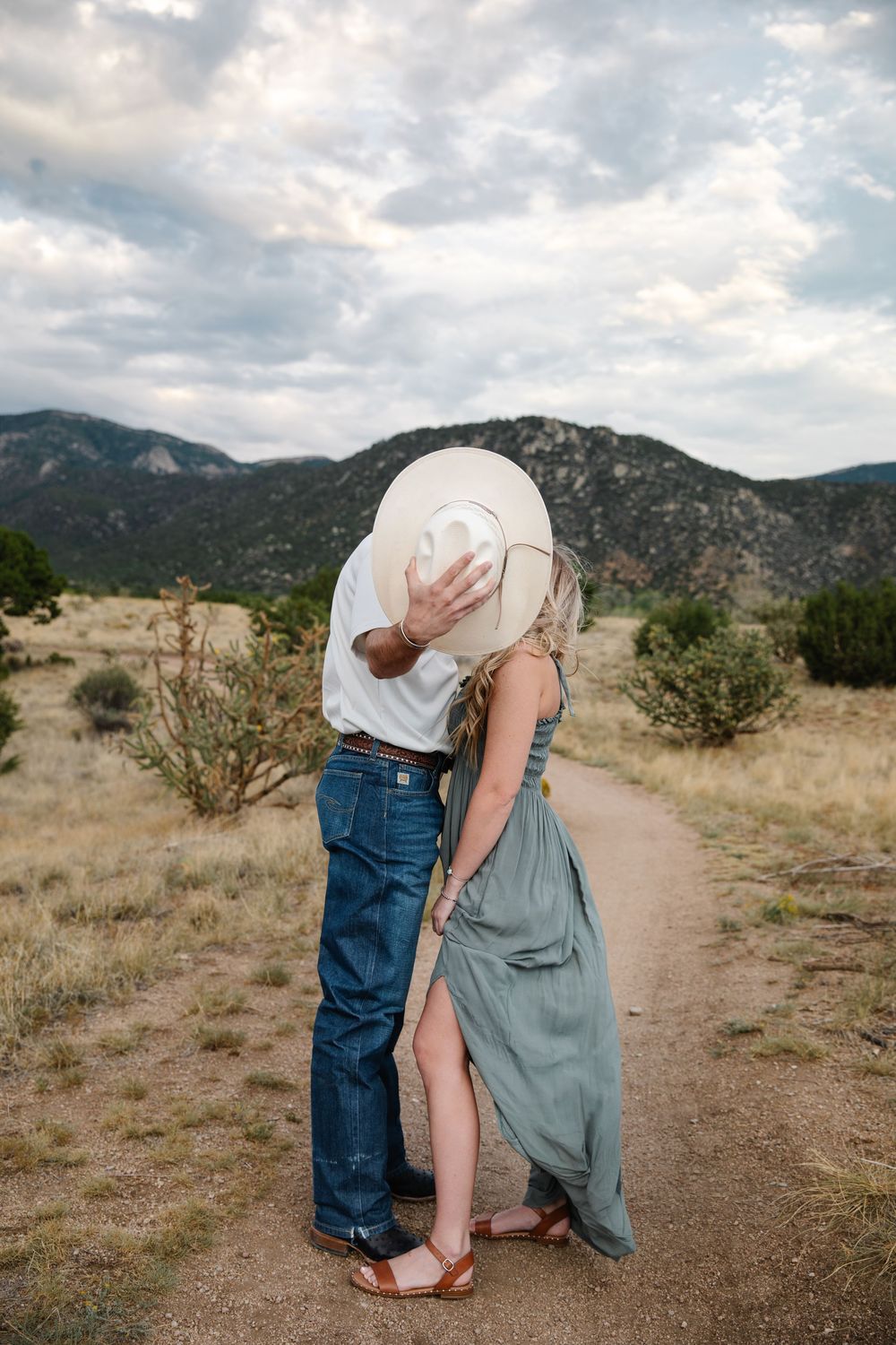 Couple kissing behind a white cowboy hat on a desert trail with mountain views.