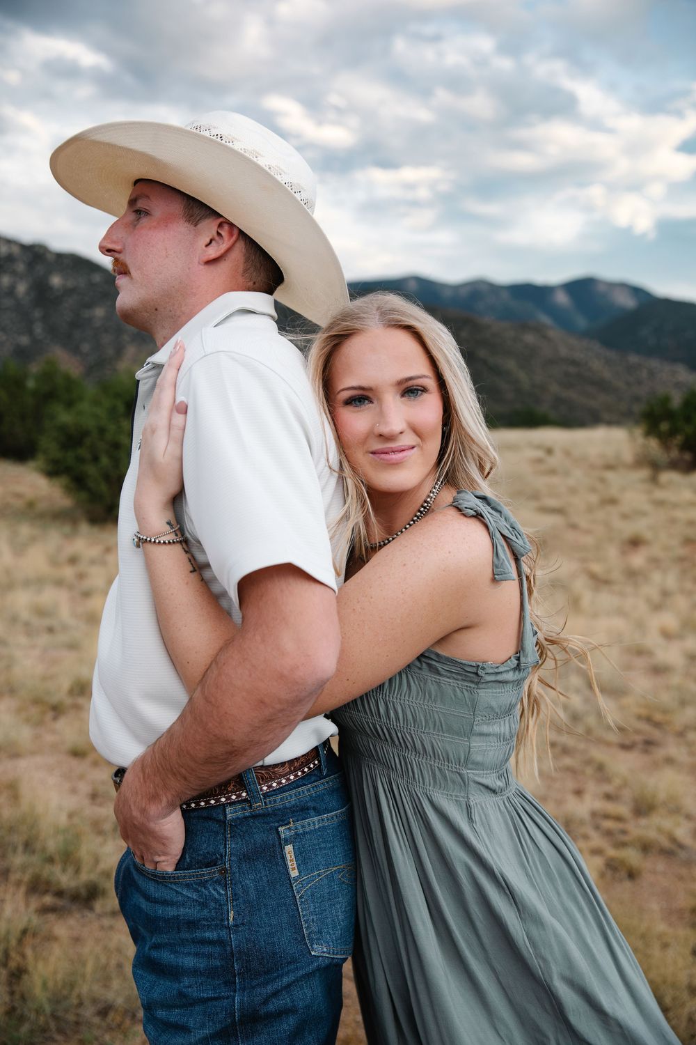 A couple embraces in a mountain meadow, woman in sage dress hugs man in cowboy hat and jeans outdoors.