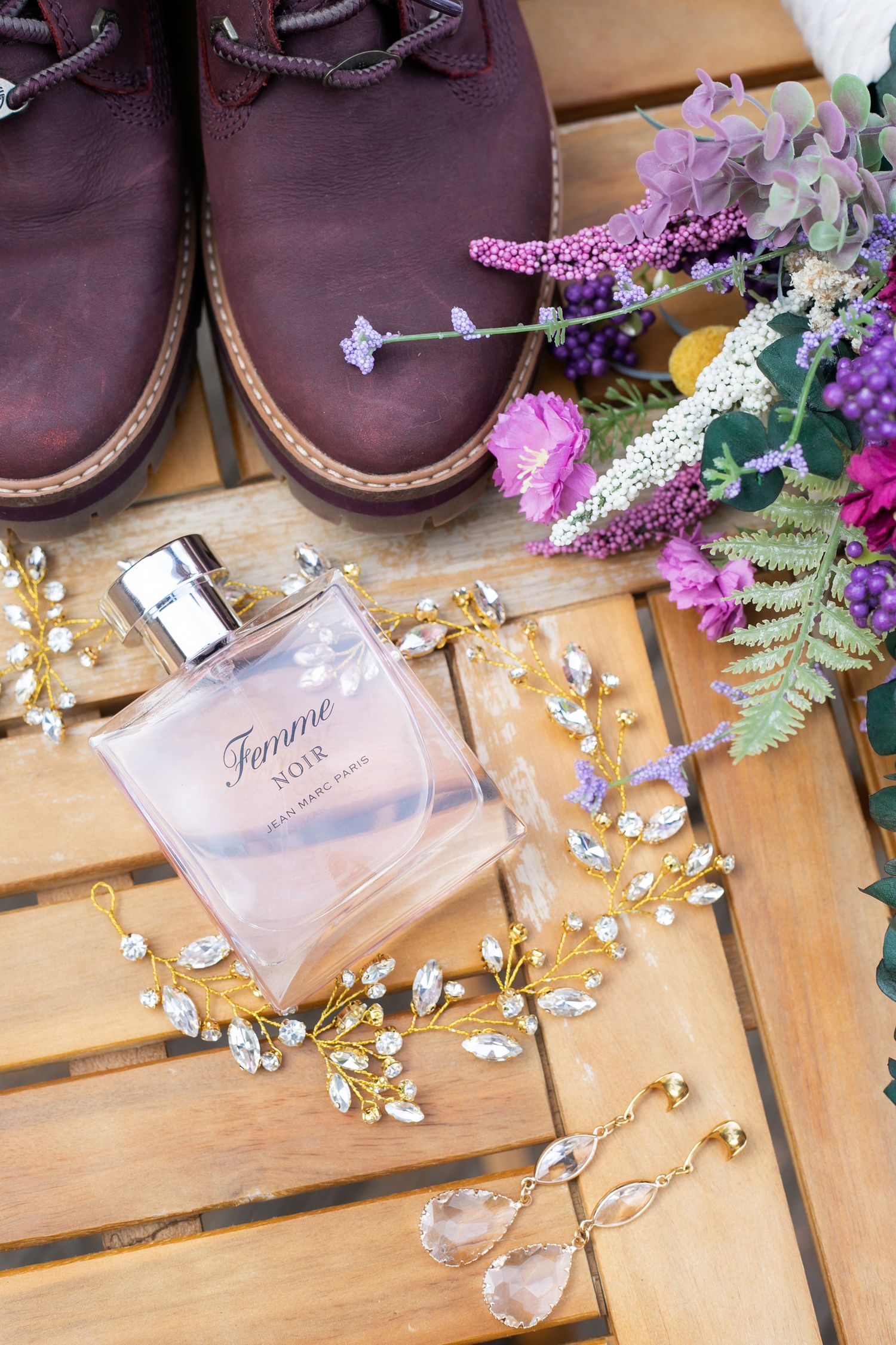 Romantic wedding details with burgundy boots, perfume bottle and purple flowers on wooden surface.