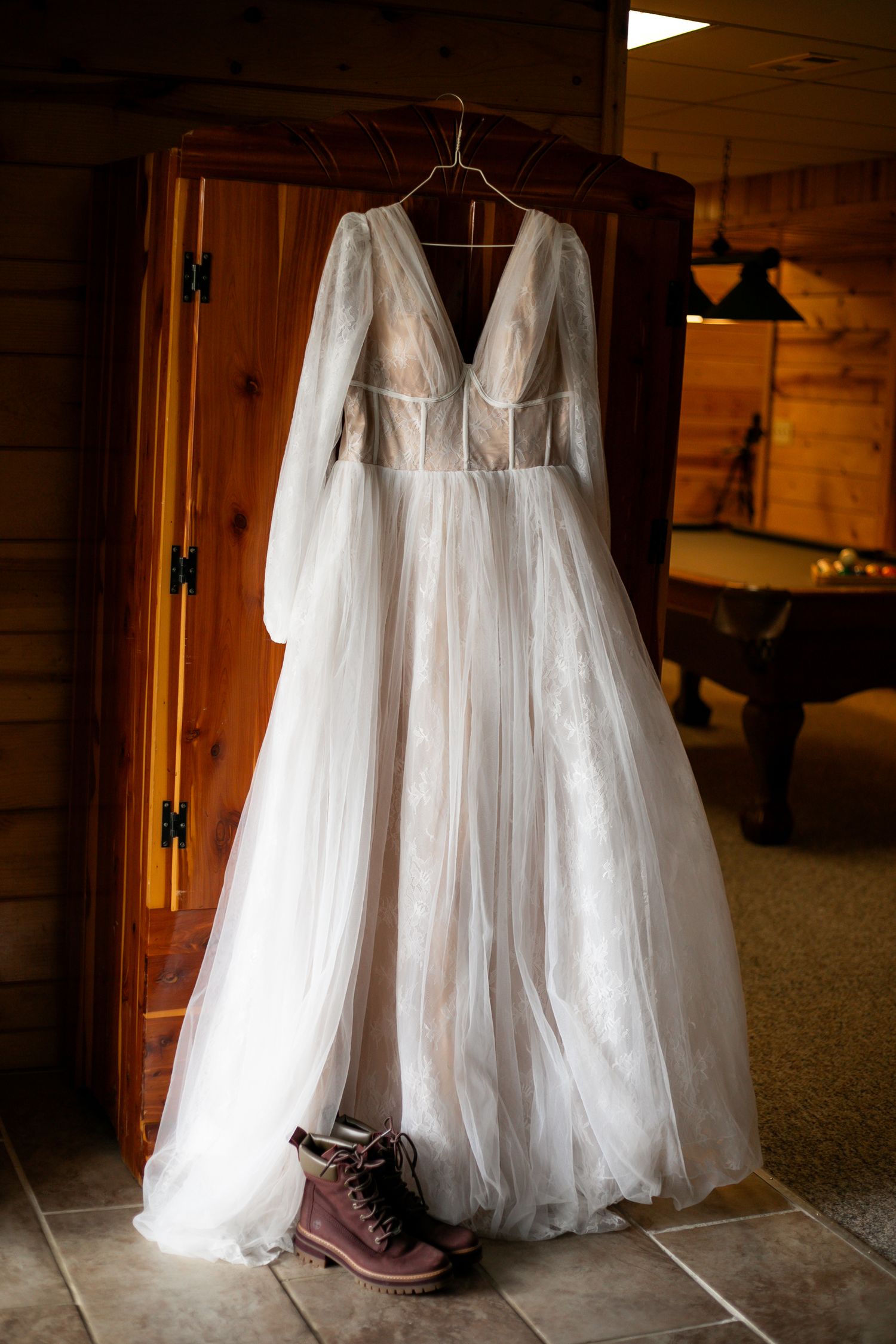 An elegant white wedding dress with long sleeves hangs beside brown boots against a wooden wall.